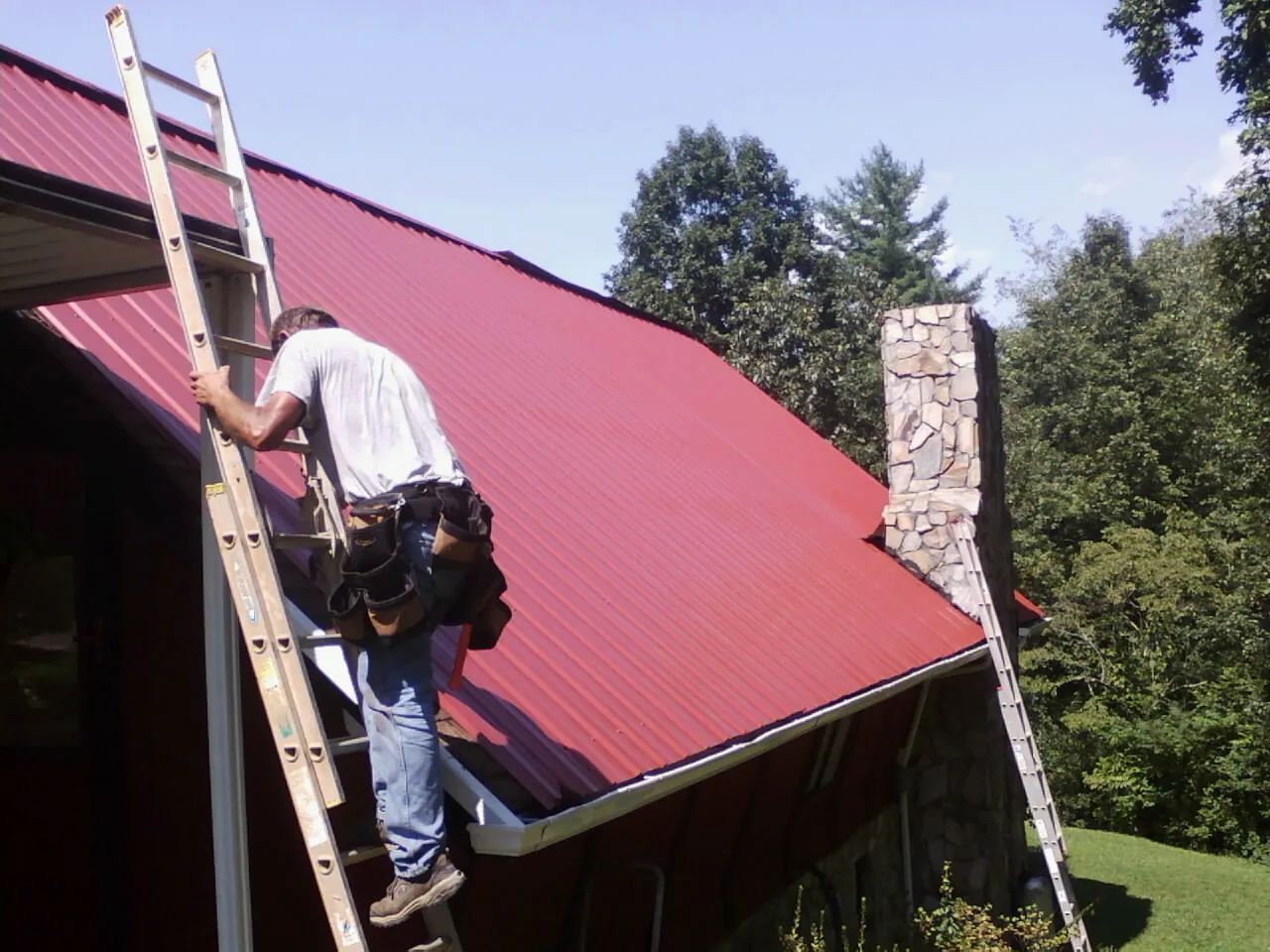 Roofer on a ladder beside a red metal roof and stone chimney — Soffit & Fascia Repair by Roofing Brothers in Bulls Gap